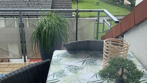 Un balcon donnant sur un quartier résidentiel, une plante en pot, une chaise en osier et une petite table recouverte d’une nappe à motifs.