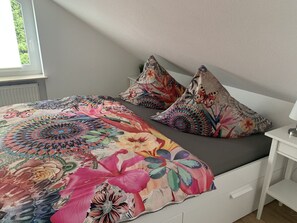 A bed with a floral-patterned bedspread and two decorative pillows, a white bedside table, and a window with a view of greenery. - Big Apartment with Lake View, Balcony and Wi-Fi (Bodman-Ludwigshafen)