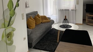 Un salon avec un canapé gris, des coussins couleur moutarde, un tapis noir, une table basse blanche et un meuble pour la télévision.
