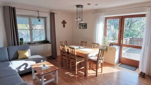 A living room with a sofa, dining table, chairs, a window with curtains, a radiator, and a cross on the wall.