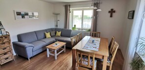 A living room with a grey sofa, wooden dining table, wicker storage, and a large window with curtains. - Holiday Apartment 'Knechtskammer' with 30m² Balcony, Shared Garden and Wi-Fi (Wilhelmsdorf)