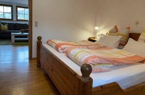A wooden bed with a striped blanket, a bedside lamp, and a view of a living room with a sofa and a window. - Holiday Apartment 'Jägerstube' with Shared Terrace, Shared Garden and Wi-Fi (Wilhelmsdorf)
