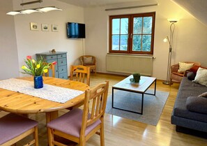 A living room with a wooden dining table, chairs, a sofa, a TV, a window with a view of trees, and a floor lamp. - Holiday Apartment 'Riedblick' with Shared Terrace, Shared Garden and Wi-Fi (Wilhelmsdorf)