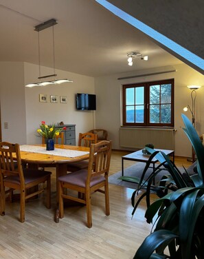 A dining area with a wooden table, chairs, a vase of flowers, a television on the wall, and a window with a view. - Holiday Apartment 'Riedblick' with Shared Terrace, Shared Garden and Wi-Fi (Wilhelmsdorf)