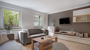 A modern living room with a sofa, coffee table, and TV. There are windows with a view of trees.