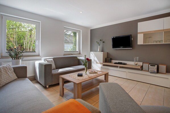 A modern living room with a sofa, coffee table, and TV. There are windows with a view of trees.