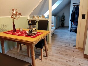 Un área de comedor con una mesa preparada para dos personas, un florero con flores secas y una ventana con vista a un edificio.