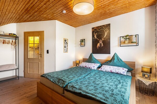 Une chambre avec un plafond en bois, un lit recouvert d’une couvre-lit à motifs verts, une tête de lit en bois, deux lampes fixées au mur et un tableau représentant un cerf, encadré.