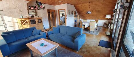 A cozy living room with a wooden ceiling, blue sofas, a small table, and a view of the kitchen.