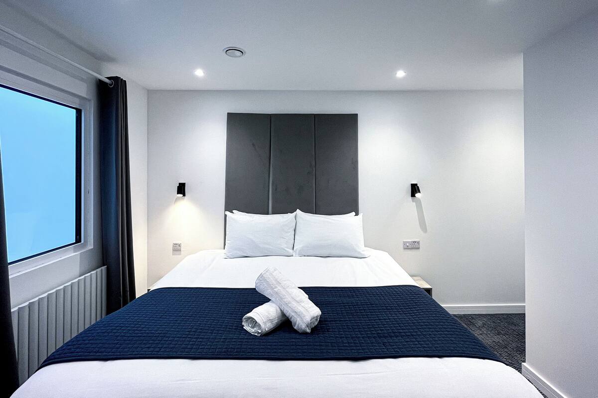 Una habitación de hotel moderna con una cama grande, una colcha azul oscuro acolchada, almohadas blancas y una toalla doblada con cuidado sobre la cama.