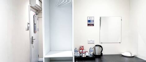 A hotel room with a shower, a desk with a kettle, a chair, and a whiteboard.