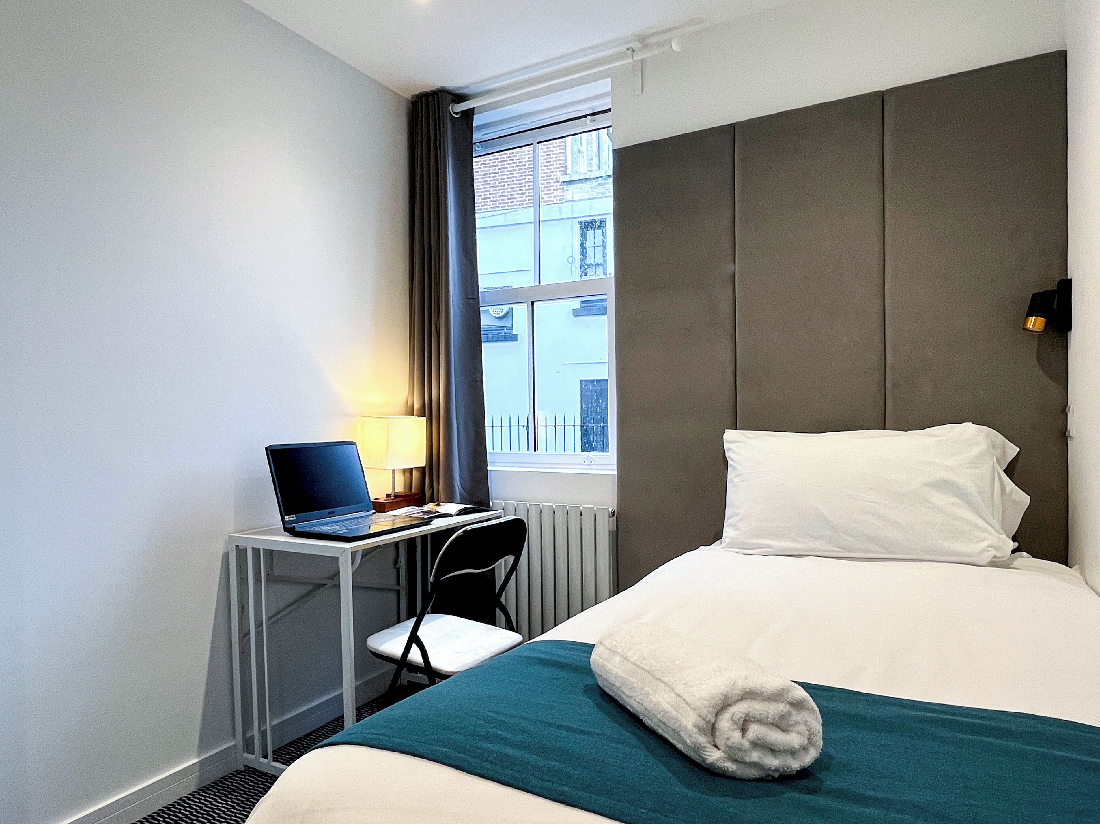 A hotel room with a bed, a desk with a laptop, a chair, and a window with a view of buildings.