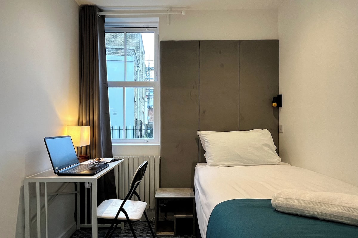 Habitación de hotel con cama, escritorio con computadora portátil, silla y ventana con vista a edificios.