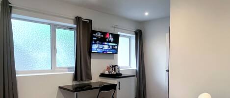 Et hotelværelse med seng, skrivebord, stol, fjernsyn monteret på væggen og et vindue med gardiner.