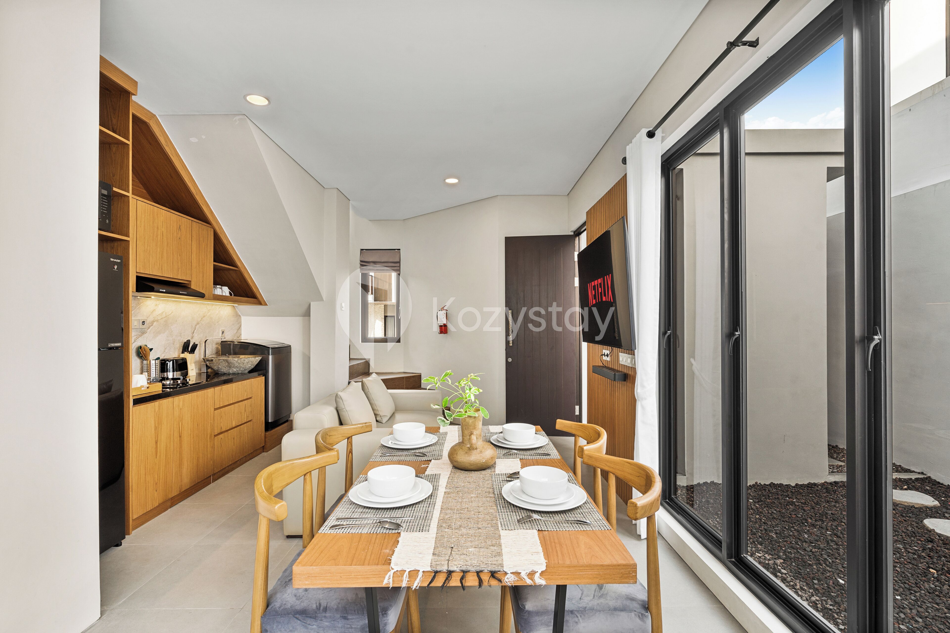 Une cuisine moderne avec des meubles en bois, un coin repas avec une table dressée pour quatre personnes, et une grande porte-fenêtre coulissante donnant sur un espace extérieur.