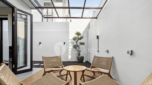 Un espacio al aire libre moderno con sillas de mimbre y una mesa redonda, rodeado de paredes blancas y un ventanal de vidrio.