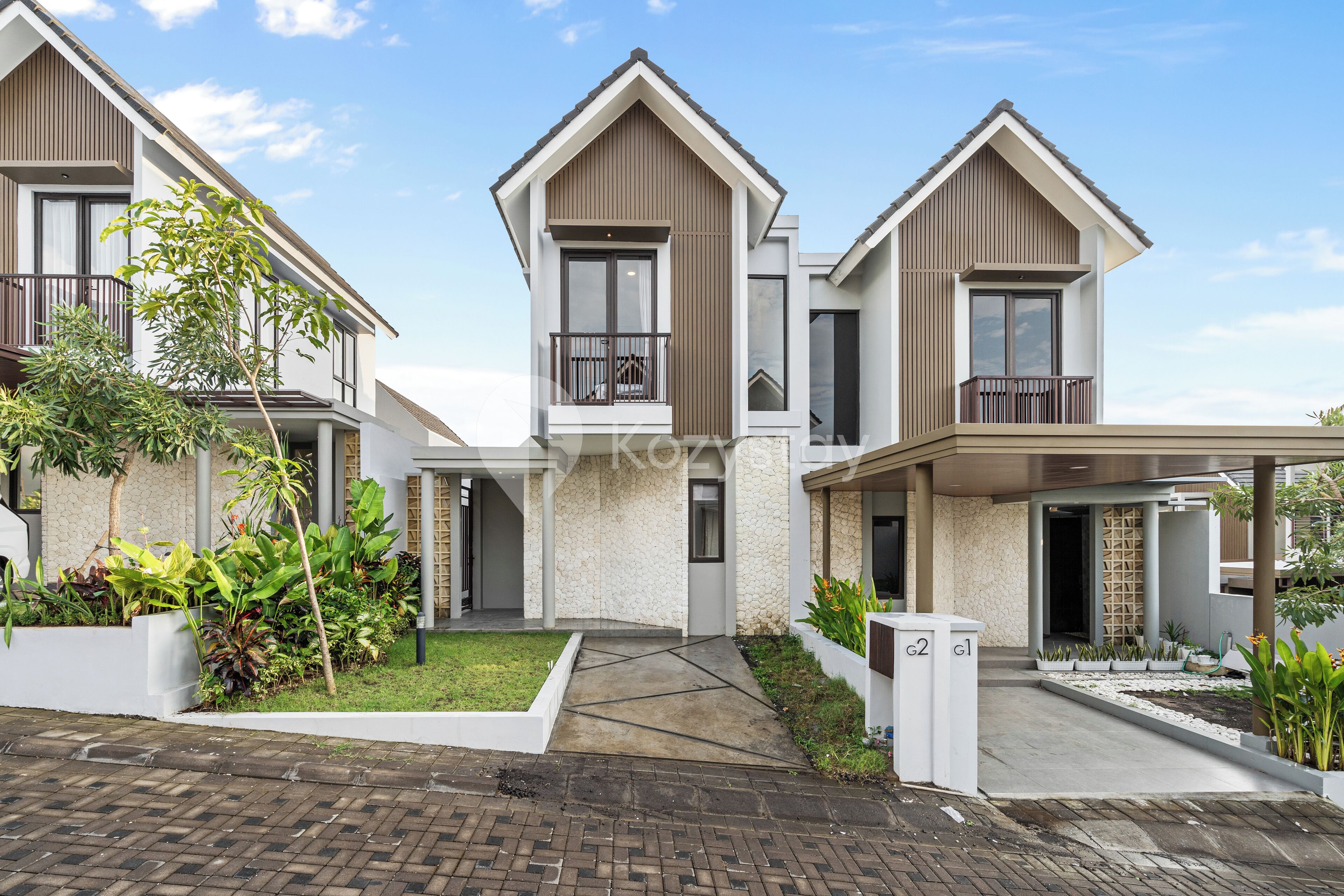A modern residential building with a symmetrical design, featuring two-story structures with balconies, a central courtyard, and a paved entrance area.