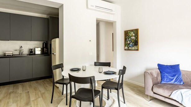 Une cuisine moderne avec des meubles de couleur sombre, une table à manger avec des chaises, un canapé avec un coussin bleu et un tableau accroché au mur.