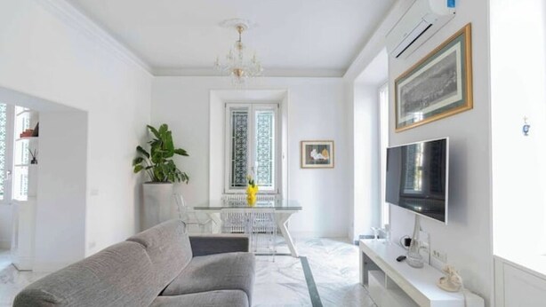 Un saló modern amb un sofà gris, una taula de menjador amb caires, un televisor penjat a la paret i una finestra gran amb cortines estampades.