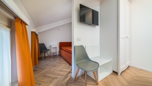 Una habitación moderna y compacta con piso de madera en patrón de espiga, televisor de pantalla plana en la pared, armario blanco y un pequeño rincón de descanso con sofá rojo y una silla verde.