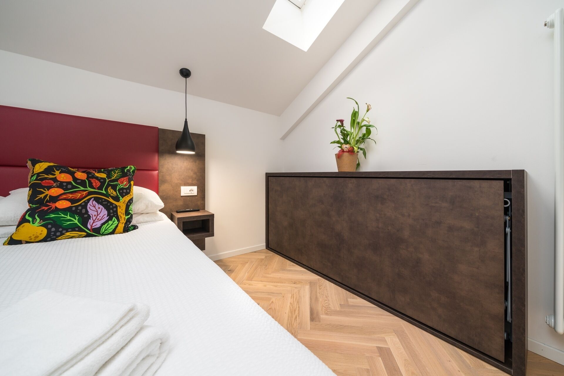 A modern bedroom with a bed, a bedside table, a nightstand, a potted plant, and a skylight.