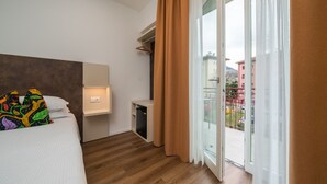 Habitación de hotel con cama, mesita de noche con lámpara, un escritorio pequeño y una puerta corrediza de vidrio que da a un balcón.