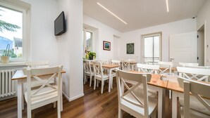 Un comedor moderno con mobiliario blanco, un televisor de pantalla plana montado en la pared y vistas al exterior a través de amplias ventanas.