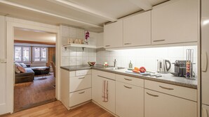A modern kitchen with white cabinets, a countertop with various appliances, and a view into a living area with a sofa and windows. - StayBrienz - Lake Brienz Holiday Apartment (Brienz)