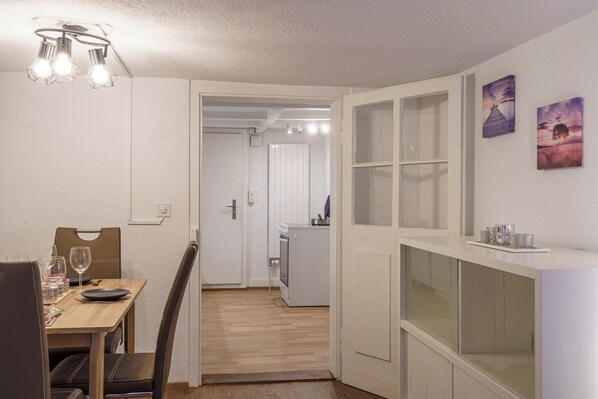 A modern kitchen with white cabinetry, a dining area with a wooden table and chairs, and a hallway leading to a bedroom.