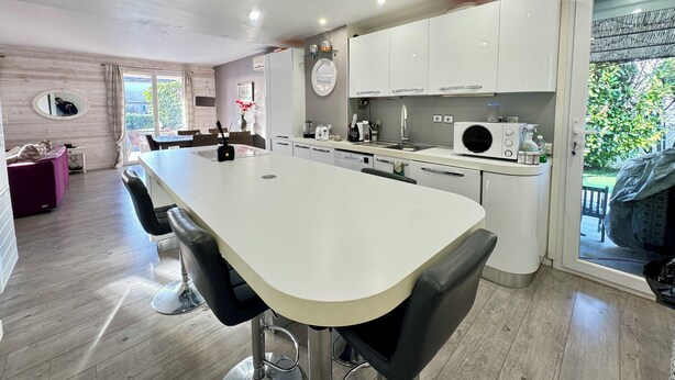 Una cocina moderna con una isla central, armarios blancos y una zona de comedor con un sofá púrpura.