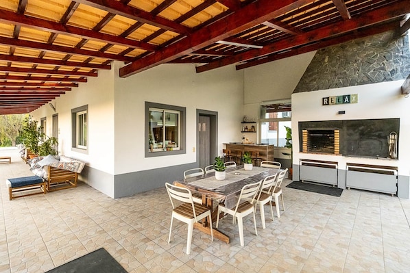 A covered outdoor dining area with a stone-tiled floor, wooden furniture, and a stone fireplace.