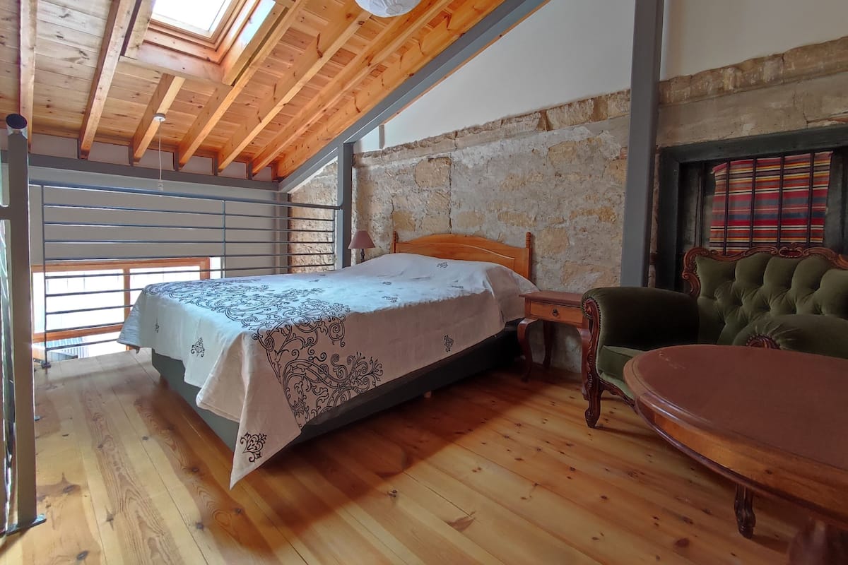 A room with a wooden ceiling, a bed with a patterned bedspread, a green armchair, and a wooden table.