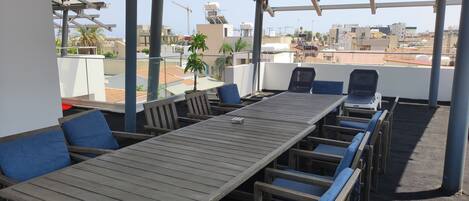Une terrasse sur le toit, avec une longue table en bois et des chaises rembourrées bleues, d’où l’on peut admirer une vue sur la ville, avec ses bâtiments et ses grues de construction.