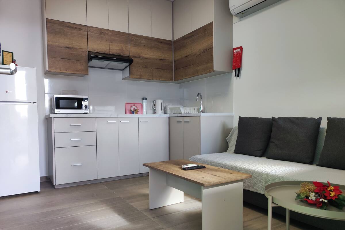 A modern kitchen with white cabinets, a microwave, and a coffee maker. There is a wooden table with a remote control and a small side table with flowers. A gray sofa with dark cushions is in the living area.