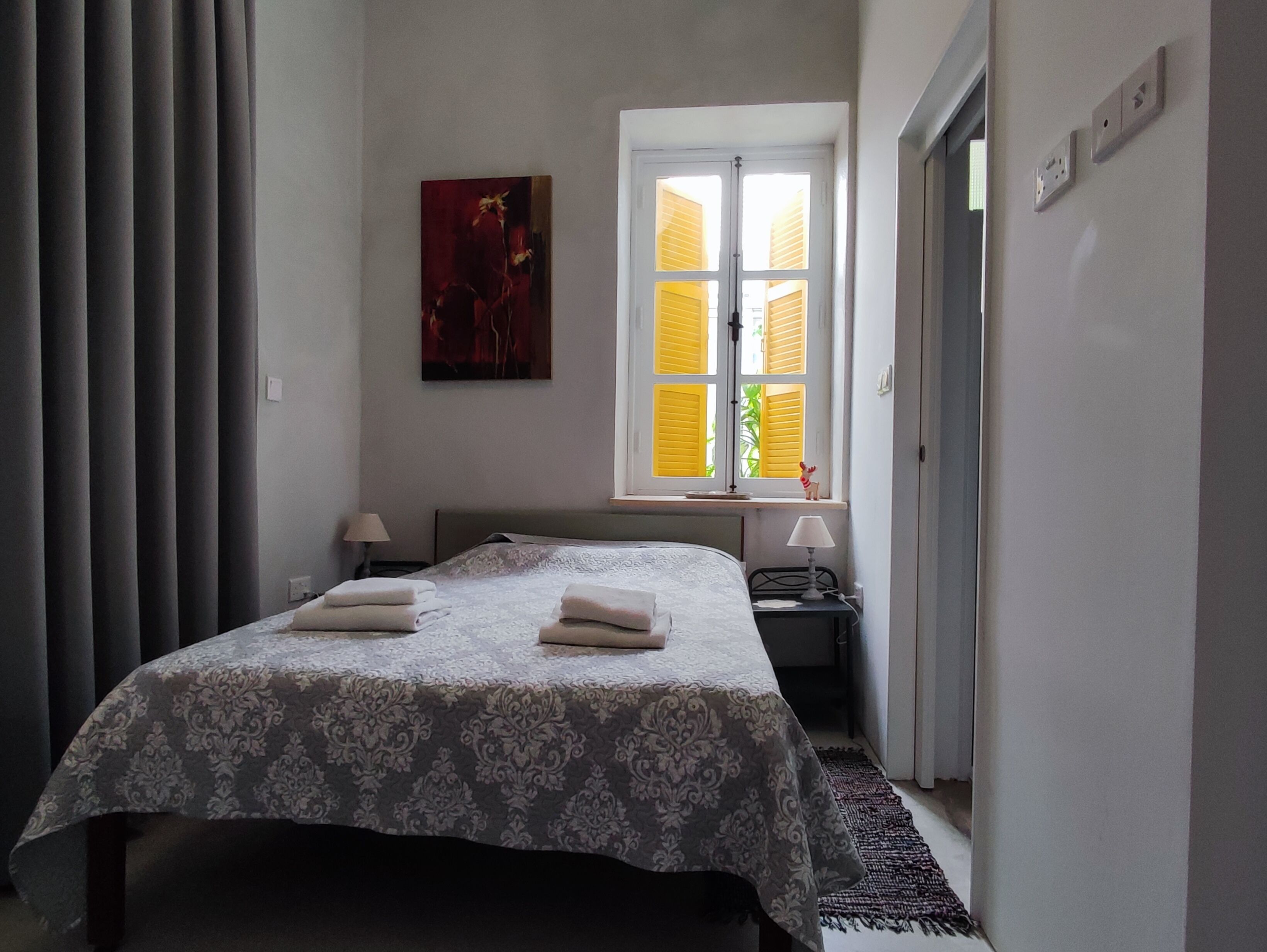 Una camera da letto con un letto, due comodini, una finestra con persiane gialle, un quadro al muro e una porta alla destra.
