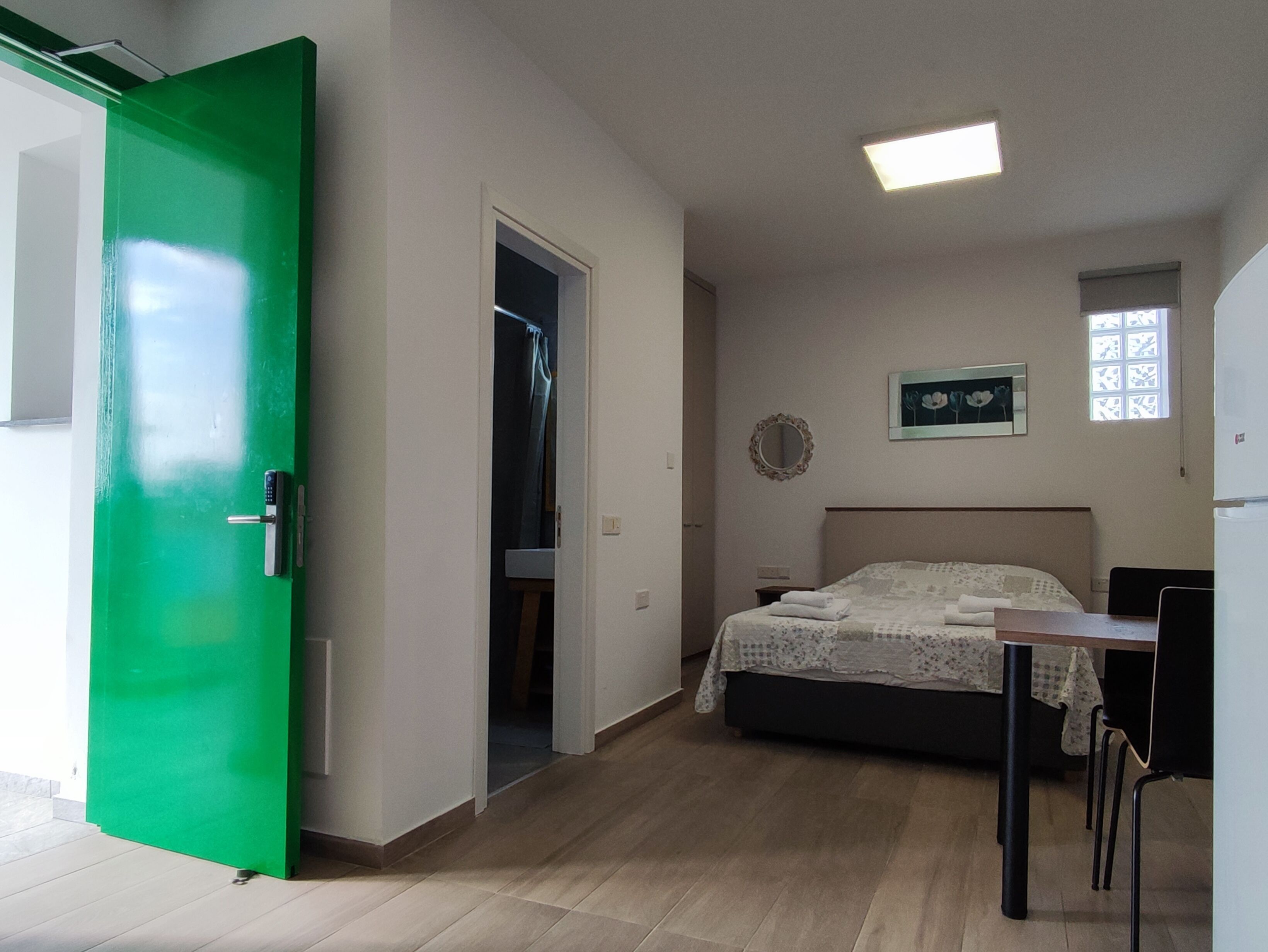 A bedroom with a bed, a desk, and a chair. There is a green door and a window with a curtain.