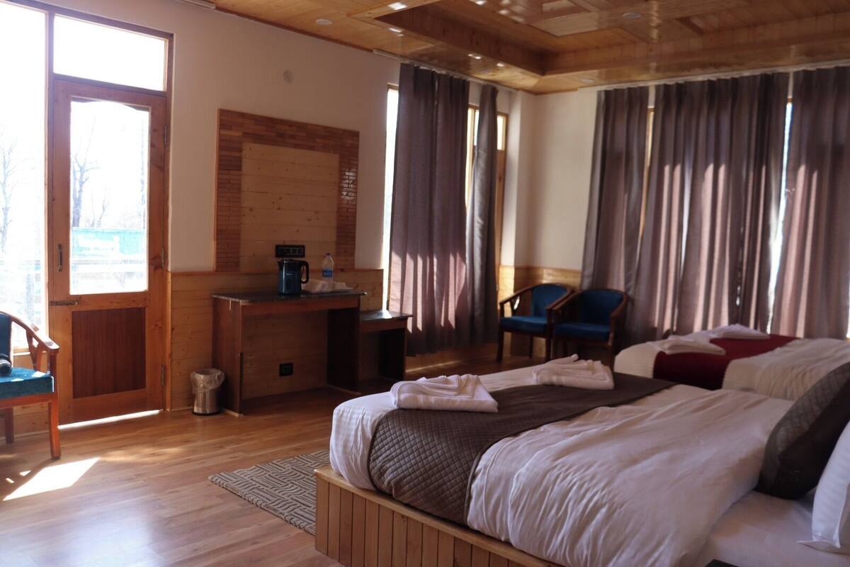 Ein Hotelzimmer mit einem hölzernen Bett, einem Schreibtisch mit Kaffeemaschine, einem Sessel und Blick ins Freie durch große Fenster.