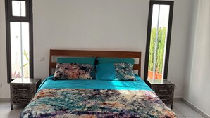 A bedroom with a wooden bed, teal bedding, and floral-patterned blankets. There are two nightstands with drawers and a window on each side. - Villa Kalélou (Nianing)