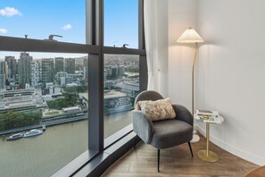 Habitación moderna con vistas a un paisaje urbano a través de amplios ventanales, un sillón gris con un cojín decorativo, una lámpara de pie y una mesita con una revista.
