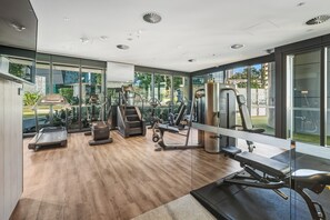 A modern gym with various exercise equipment, large windows, and a wooden floor.
