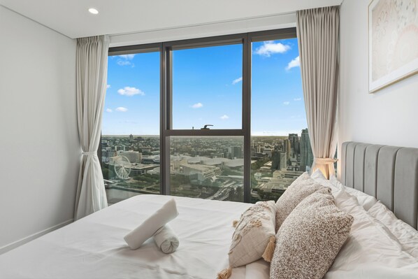A hotel room with a large window offering a city view, a neatly made bed with pillows, and a headboard.