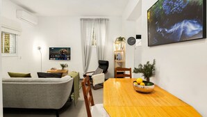 Ein modernes Wohnzimmer mit einem grauen Sofa, einem Holzesstisch mit Obstschale und einem großen Fernsehbildschirm, der eine Naturlandschaft zeigt.