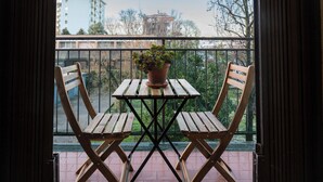 Un balcón con una mesa de madera y dos sillas, una planta en maceta sobre la mesa, y vistas a edificios y árboles al fondo.