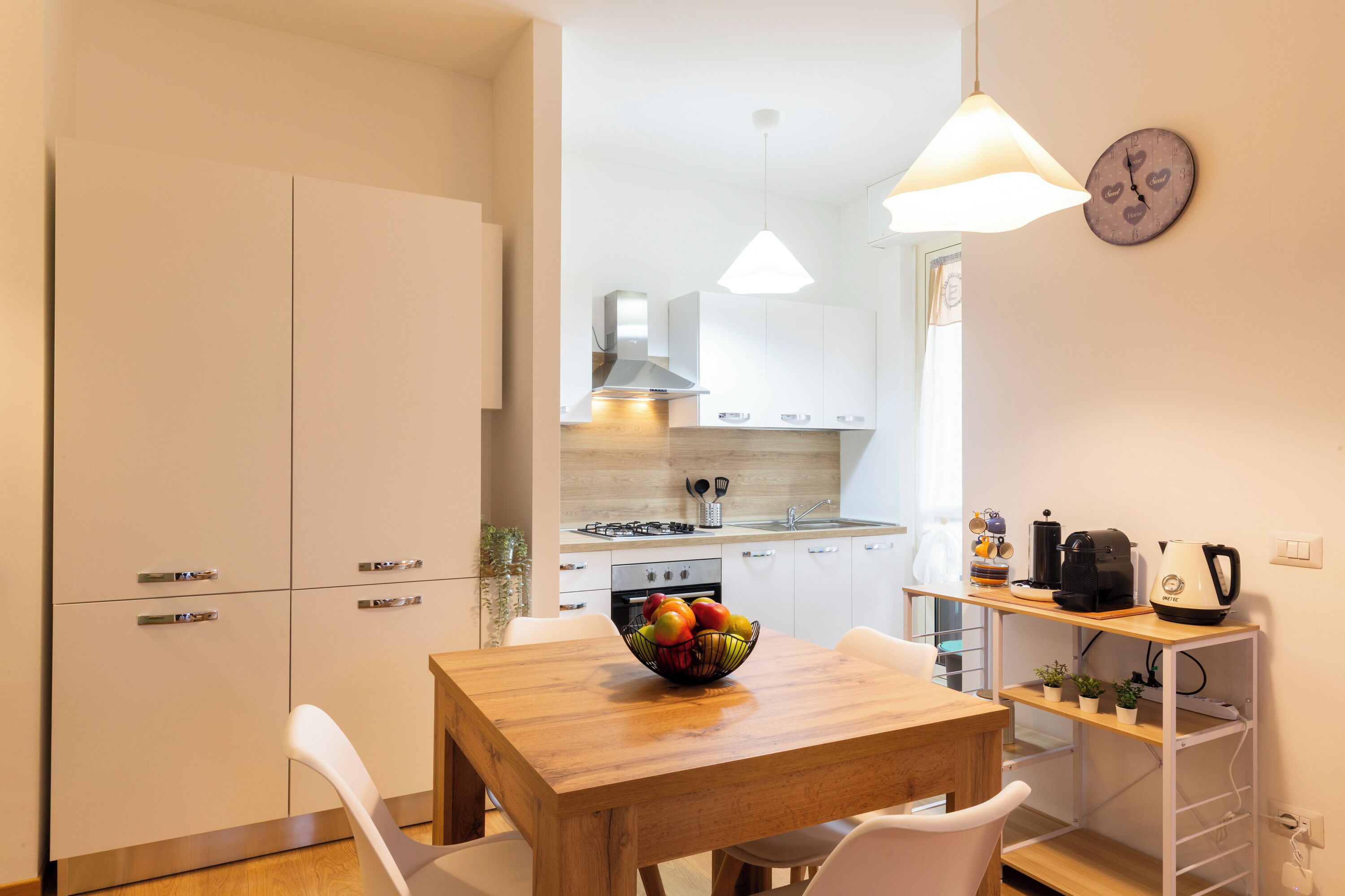 A modern kitchen with white cabinets, a wooden dining table, and a bowl of fruit.