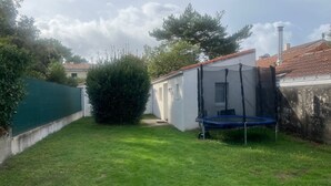 Un patio trasero pequeño y cercado con un trampolín, un edificio blanco de techo rojo y una valla verde.