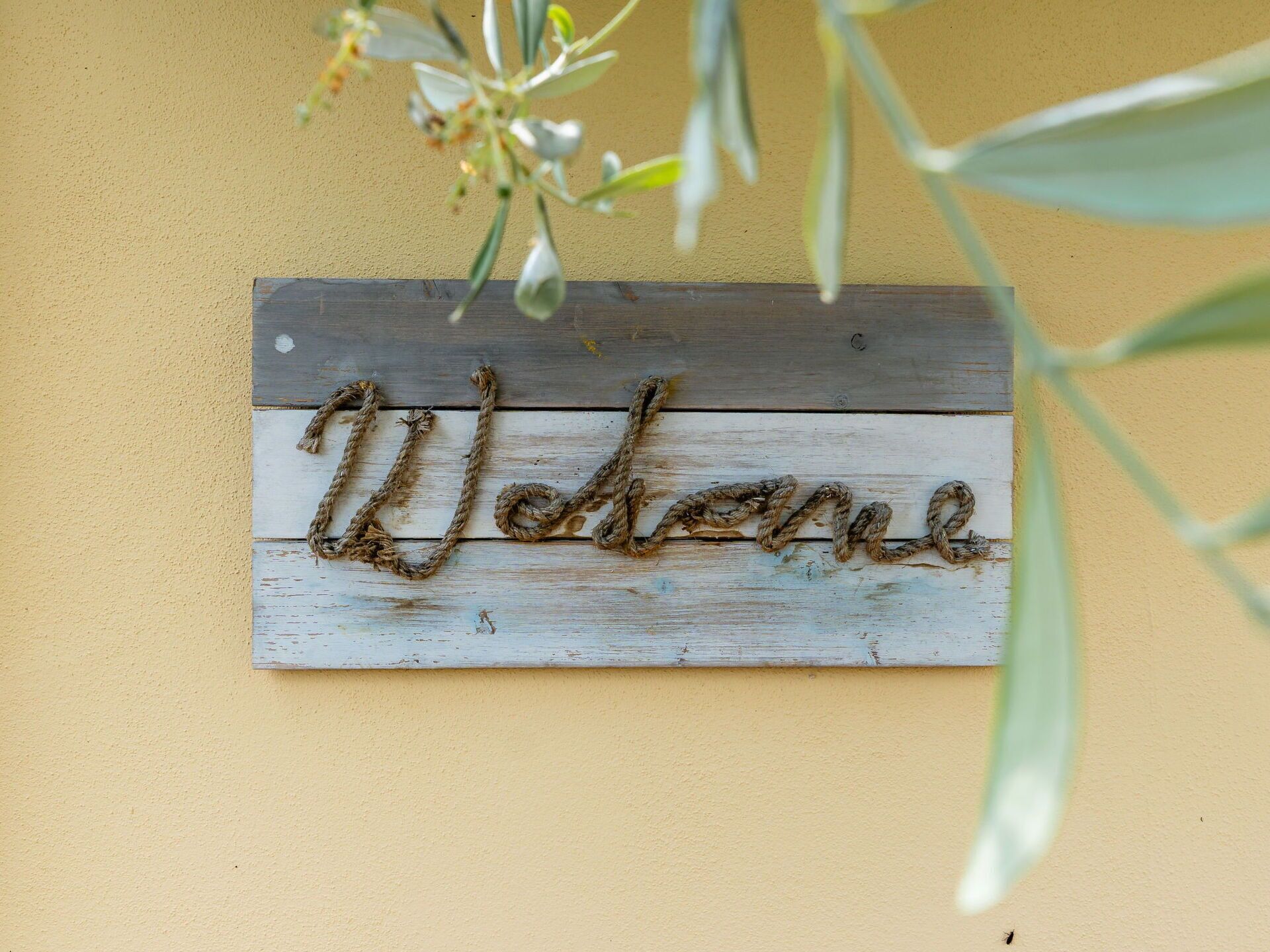A rustic wooden sign with the word "Welcome" written in a cursive, weathered font, mounted on a wall with a textured yellow surface.
