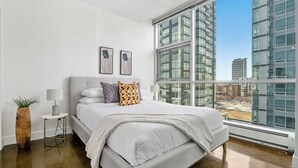 A modern bedroom with a large window, a bed with white bedding, a side table with a lamp, and two framed pictures on the wall.
