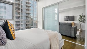 A modern hotel room with a large bed, a grey sofa, a TV, and a view of a high-rise building.
