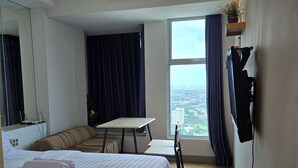 A hotel room with a bed, a small table, a chair, a TV mounted on the wall, and a large window offering a city view. - Cosmy Benson 38 Apartment (Surabaya)