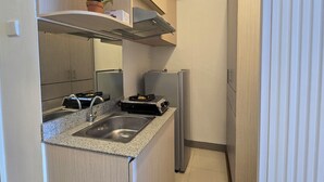 A compact kitchen with a built-in stove, sink, and a small refrigerator. There are wooden cabinets and a potted plant on a shelf. - Cosmy Benson 38 Apartment (Surabaya)